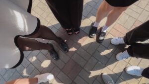 A group of individuals stands in a circle on sunlit paved ground. Only their feet and legs are visible, showcasing a variety of stylish footwear, including black heels, loafers, and white sneakers. The ground features a gray and brown brick pattern.